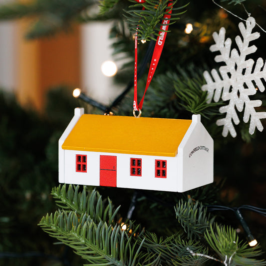 A small house-shaped Christmas ornament hanging on a green Christmas tree. The ornament is white with a yellow-orange roof, red door and window details, and a red hanging ribbon.