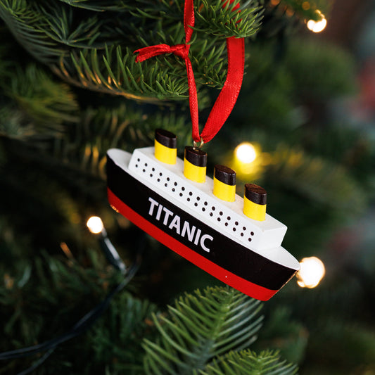 A miniature Titanic Christmas ornament hanging from a red ribbon on a green Christmas tree with warm string lights.