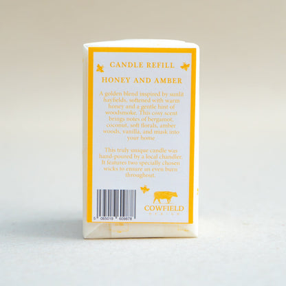 A candle refill package standing upright on a light neutral background. The front is mostly obscured by a white rectangular overlay, but the visible side and back panels are cream with yellow border lines and text reading 'CANDLE REFILL' and 'HONEY AND AMBER,' plus the Cowfield brand name and logo.