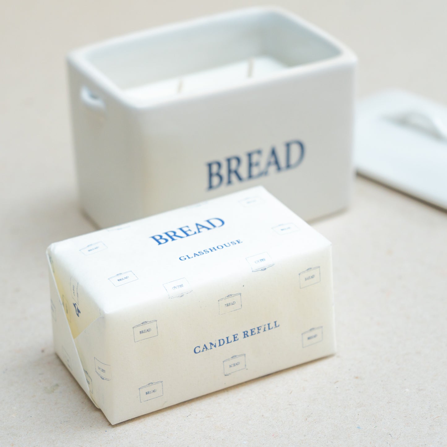 A candle refill package labeled 'BREAD' in blue text on an off-white wrapper, with the matching white candle container and lid blurred in the background.