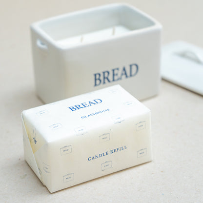 A candle refill package labeled 'BREAD' in blue text on an off-white wrapper, with the matching white candle container and lid blurred in the background.