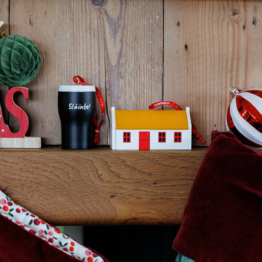 A styled holiday shelf scene with a small white house ornament featuring a yellow roof and red trim, next to a black pint with red ribbon , against a rustic wood plank background.