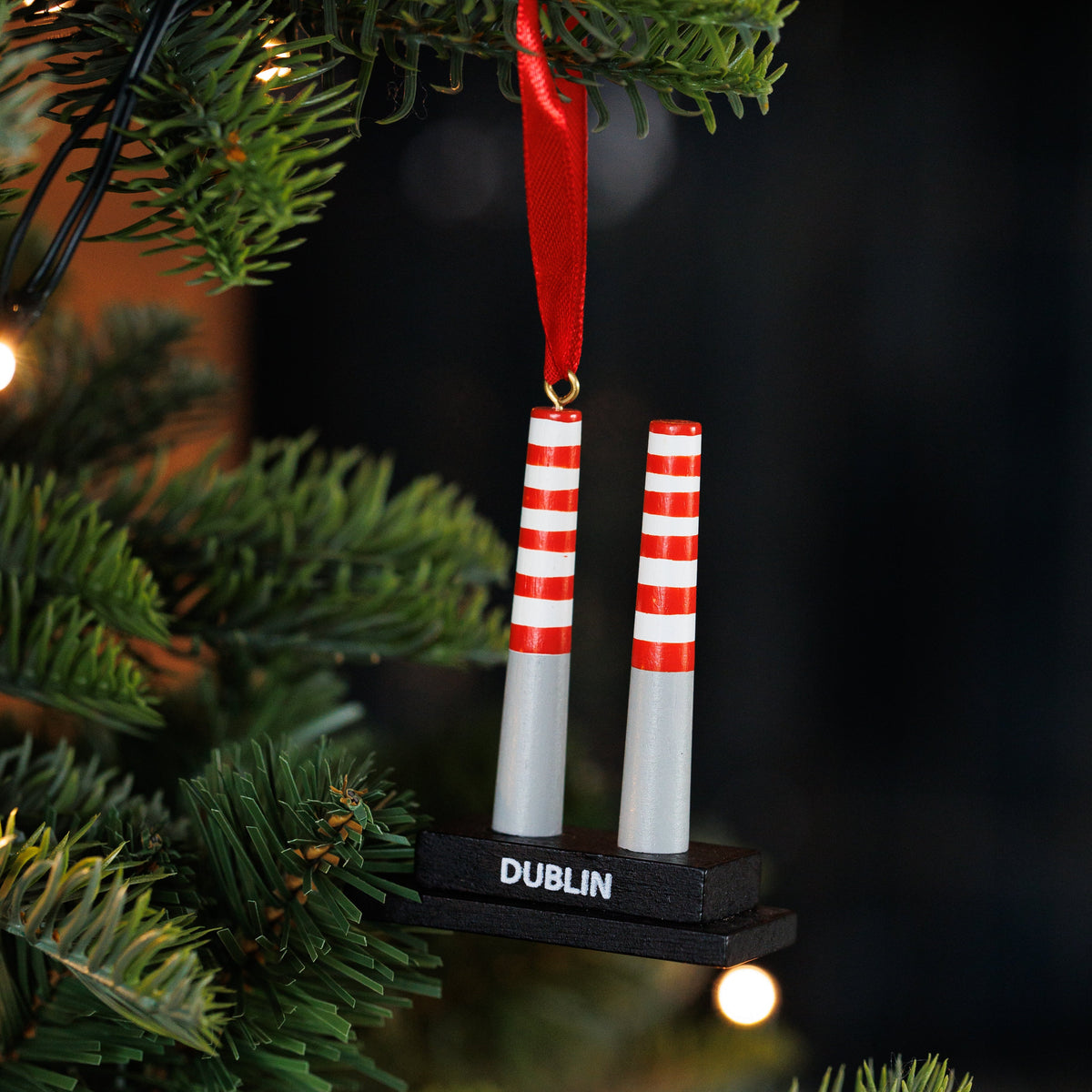A Christmas ornament shaped like poolbeg towers with red and white striped signs on a black base, hanging from a green Christmas tree with warm string lights.
